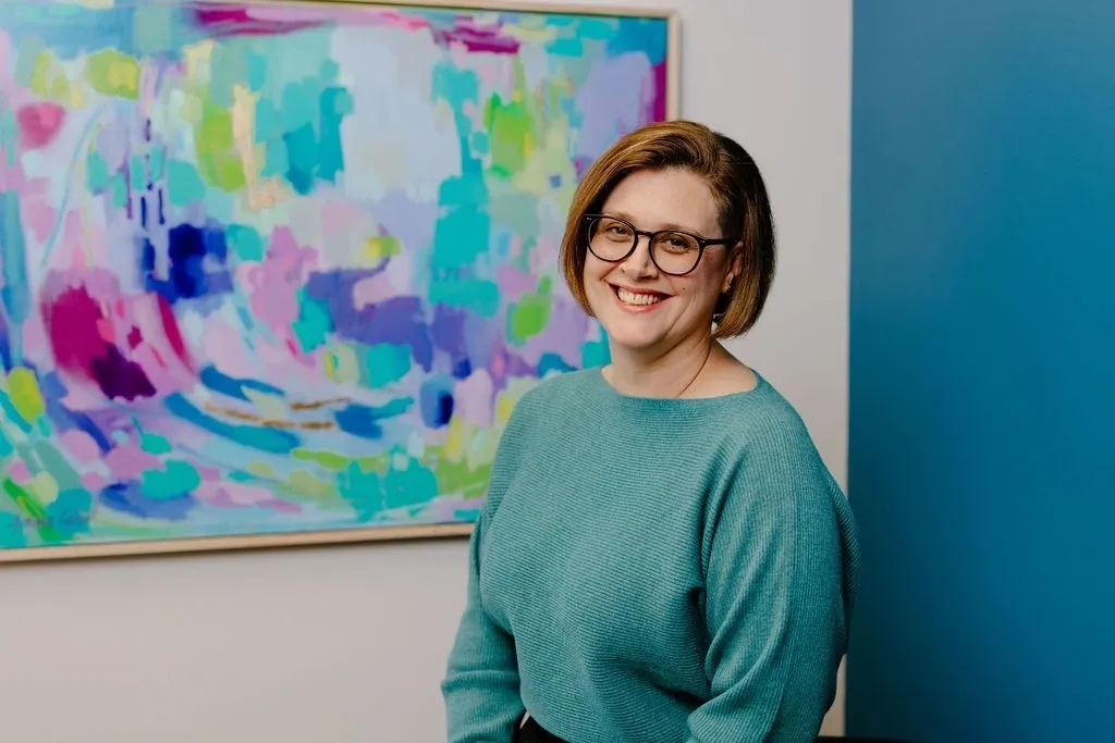 Woman with glasses smiling in front of colorful abstract painting.