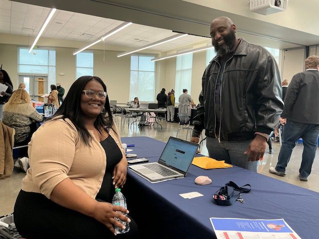 Two people smiling at a professional event with laptops and informational materials.