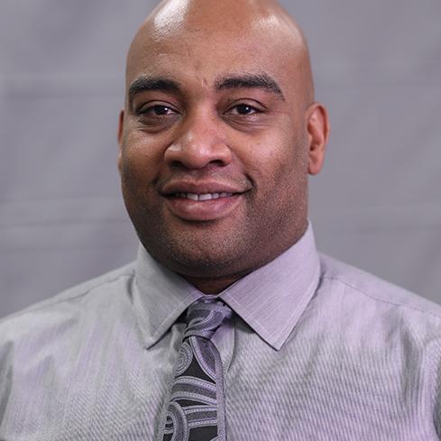 Portrait of a smiling man in a gray shirt and patterned tie.