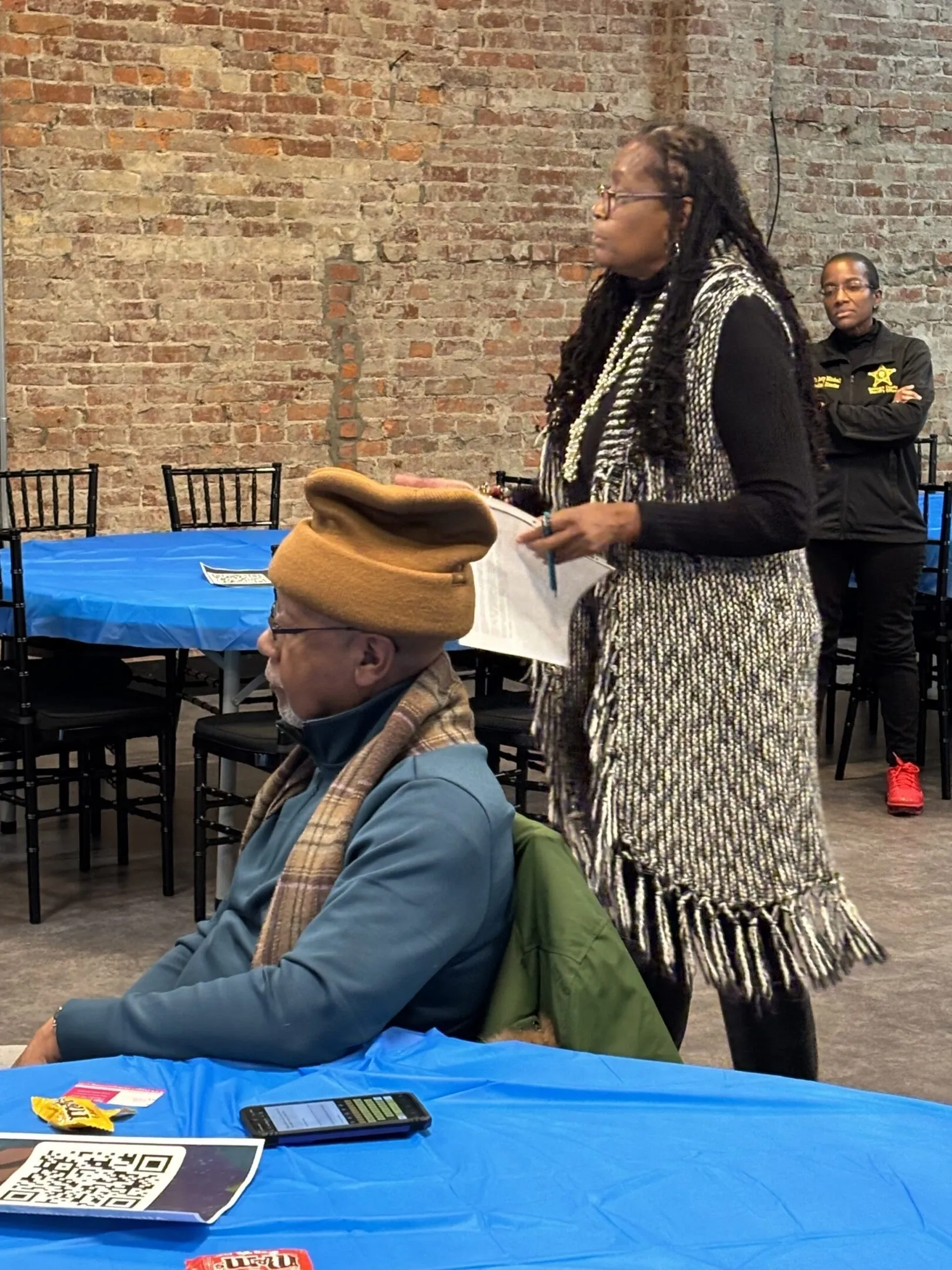 A seated man in a tan hat and scarf listens to a standing woman speaking at an event.