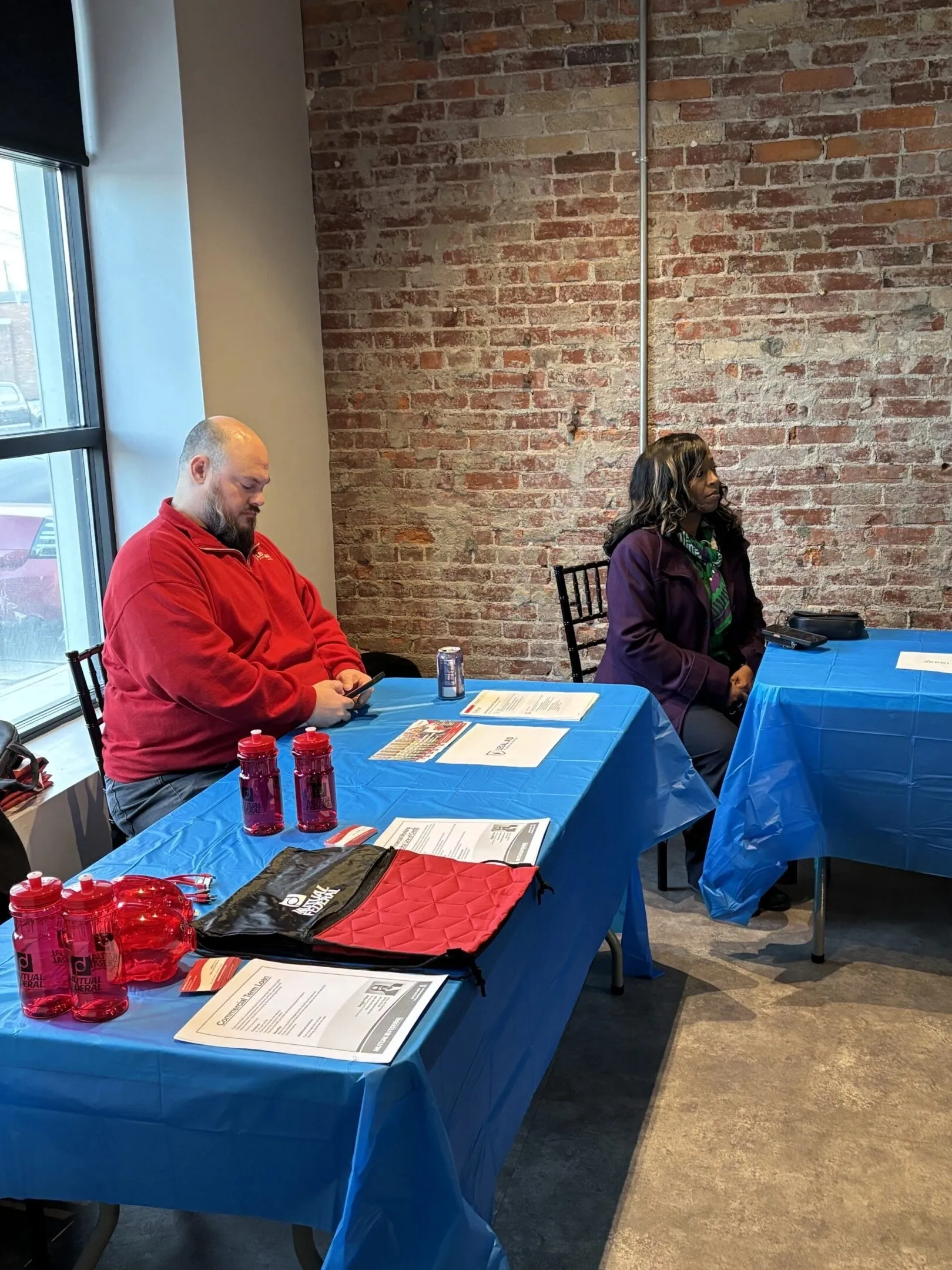 Two people sitting at separate tables covered with blue cloths inside a brick-walled room.