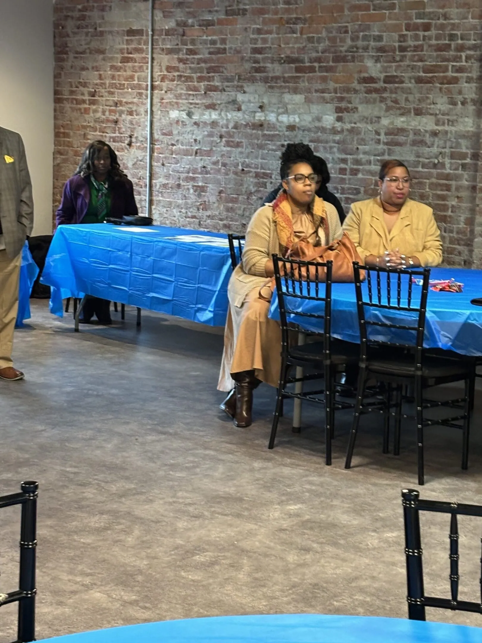 Two women sitting at a table with blue tablecloth in a room with brick walls.